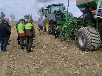 Foto: Abgeerntetes Maisfeld. Linker Hand eine Gruppe von Landwirten schaut auf ein Gespann aus Traktor und einer Direktsaatmaschine. Ein Mann hockt vor der Maschine und erklärt etwas.