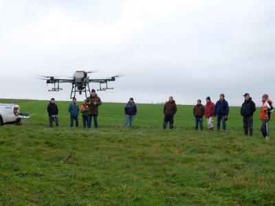 Foto: auf einem Acker stehen im Hintergrund Menschen und schauen auf eine Drohne, die im Vordegrund fliegt
